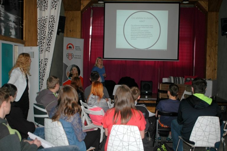 Grupa młodych os&oacute;b siedzi w sali z ekranem. Na ekranie widać napis czy właśnie to chcę robić? W tle stoi baner Centrum Zimbardo.