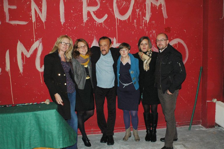 Profesor Zimbardo w towarzystwie czterech kobiet i mężczyzny pozuje do zdjęcia na tle czerwonej ściany z białym napisem centrum.