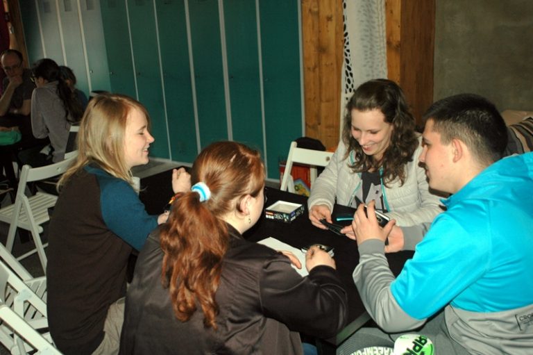 Czworo młodych ludzi siedzi przy czarnym stole i gra w grę karcianą. W tle widać turkusowe szafy.