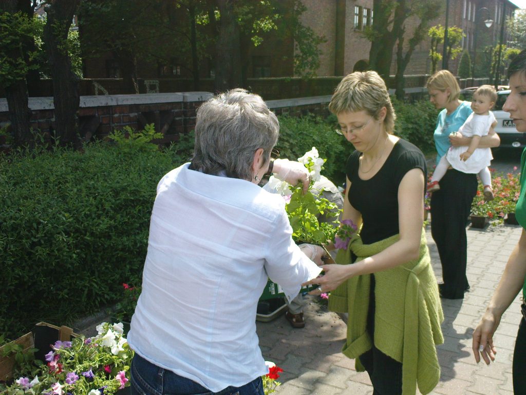 Dwie kobiety przekazują sobie sadzonki kwiat&oacute;w na chodniku W tle kobieta z małym dzieckiem na ręku.