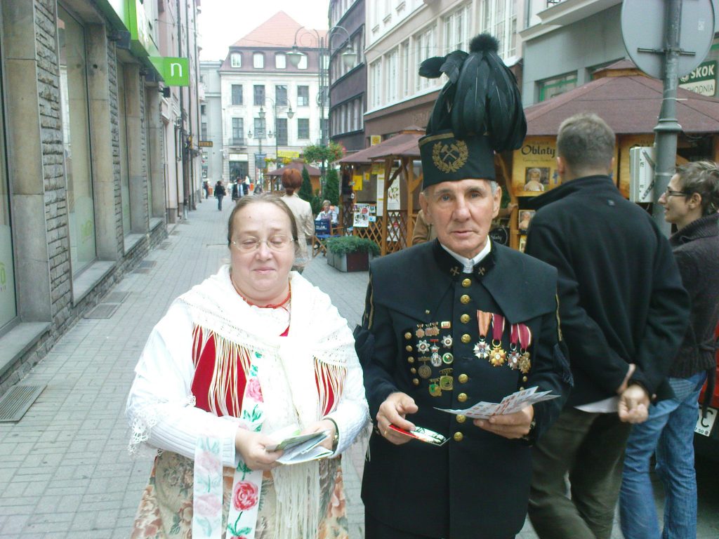 Kobieta w śląskim stroju ludowym i mężczyzna w mundurze g&oacute;rniczym idą ulicą, trzymają w ręku ulotki. W tle kamienice.