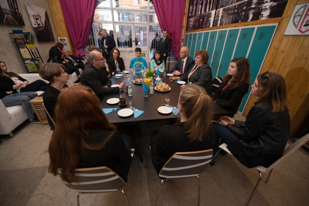 Uczestnicy spotkania z Anną Komorowską. Osoby siedzą przy stole. W tle ochroniarze Pierwszej Damy i reporterzy.