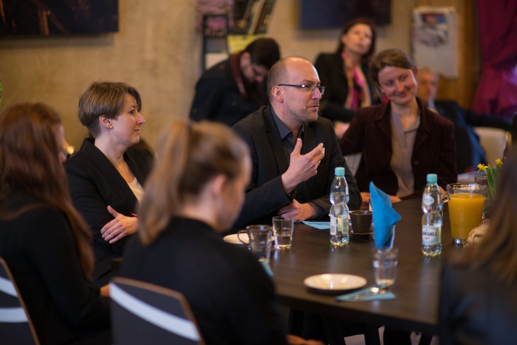 Uczestnicy spotkania siedzą przy stole. Na gł&oacute;wnym planie mężczyzna w okularach gestykulujący w trakcie rozmowy.