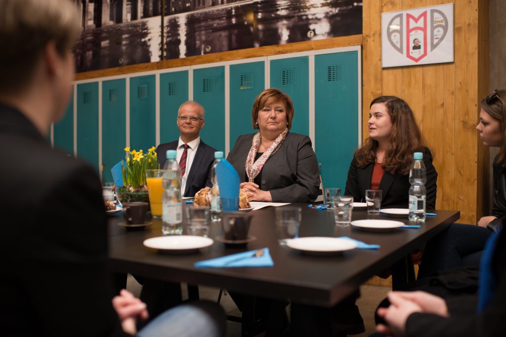 Pierwsza dama z przedstawicielami Centrum Zimbardo siedzi przy stole, w tle turkusowe szafki i logo Centrum Zimbardo wiszące na ścianie.