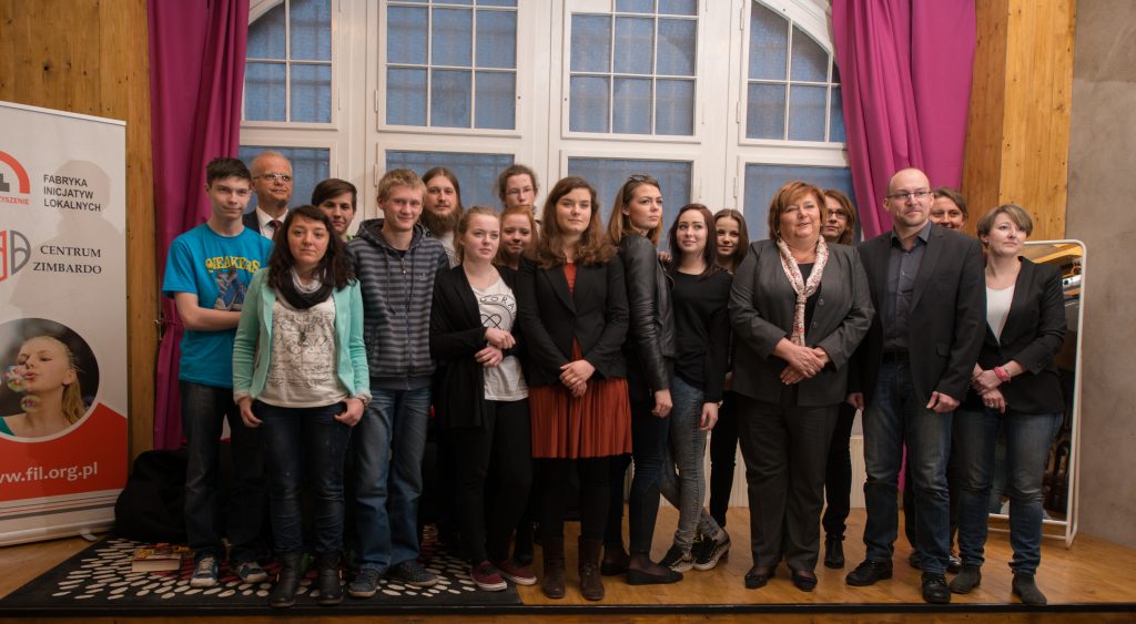 Anna Komorowska z przedstawicielami Centrum Zimbardo w W Katowicach-Nikiszowcu. Osoby stoją na scenie, obok baner z logotypami Fabryki Inicjatyw Lokalnych i Centrum Zimbardo.