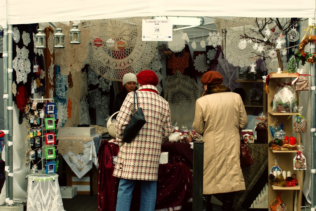 Dwie kobiety rozmawiają z osobą przy stoisku z serwetami robionymi szydełkiem.