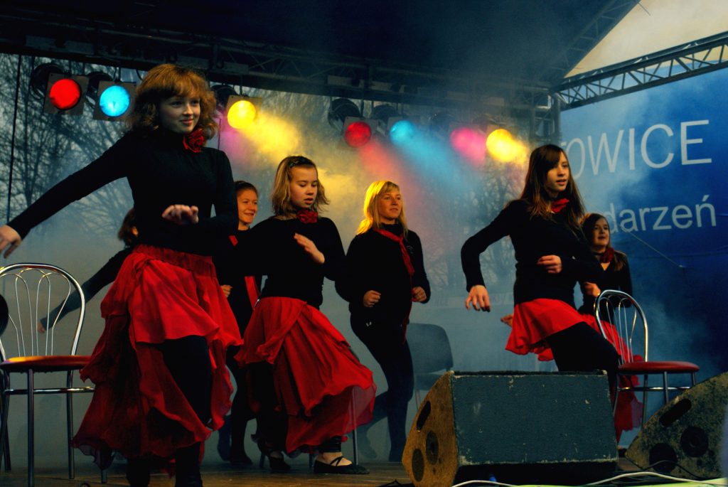 Grupa dziewcząt ubranych w czarne bluzki i czerwone sp&oacute;dnice tańczy na scenie.