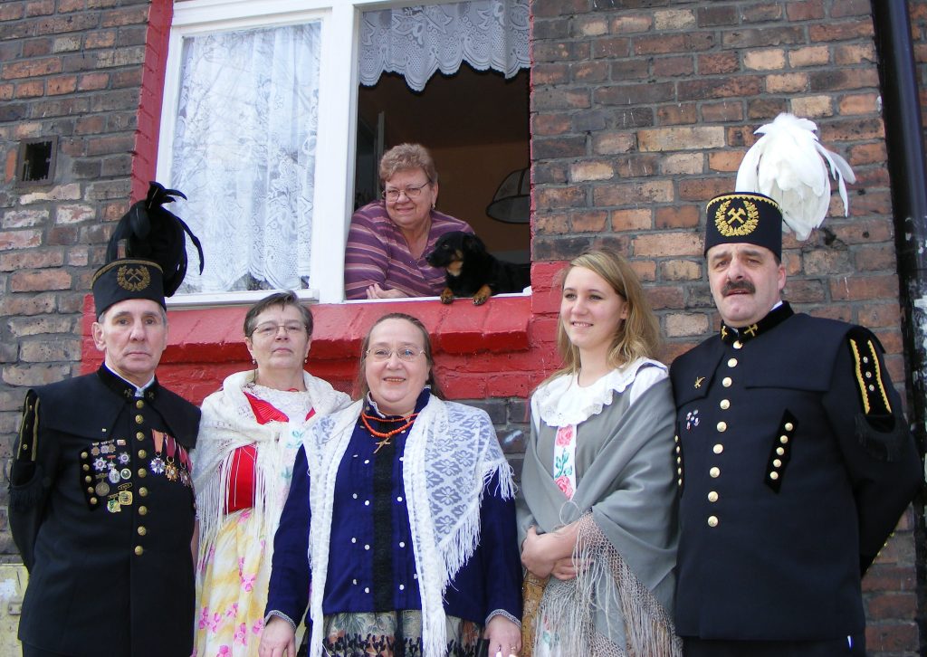 Grupa os&oacute;b w śląskich strojach regionalnych i g&oacute;rniczych mundurach galowych pozuje przed ceglanym budynkiem. W otwartym oknie nad nimi widać uśmiechniętą kobietę z małym psem.