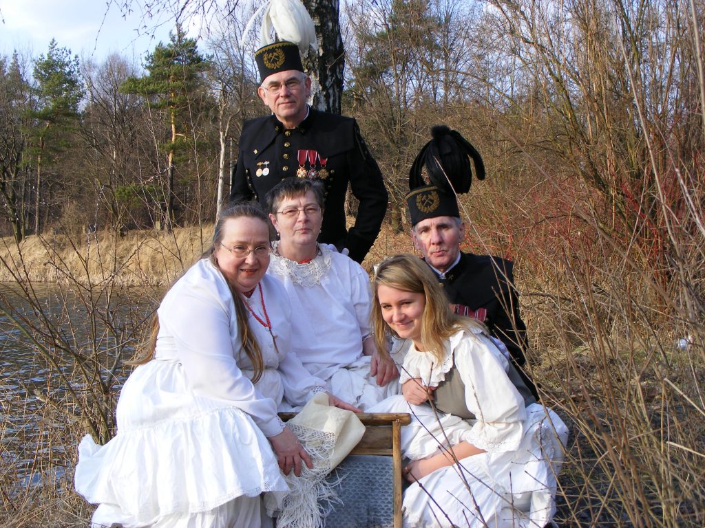 Dwaj mężczyźni w galowych mundurach g&oacute;rniczych i trzy kobiety w białych, tradycyjnych sukniach, trzymające dawną tarkę do prania. Osoby pozują nad wodą.