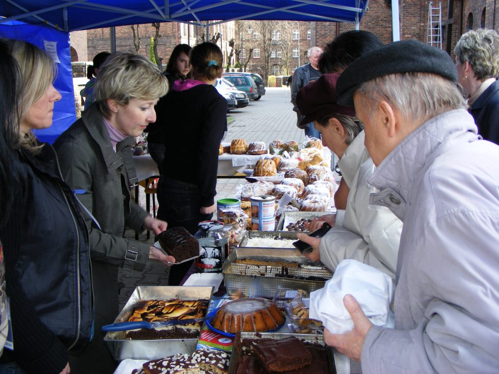 Grupa os&oacute;b przy stoisku z domowymi wypiekami ustawionymi na stole pod niebieskim namiotem.