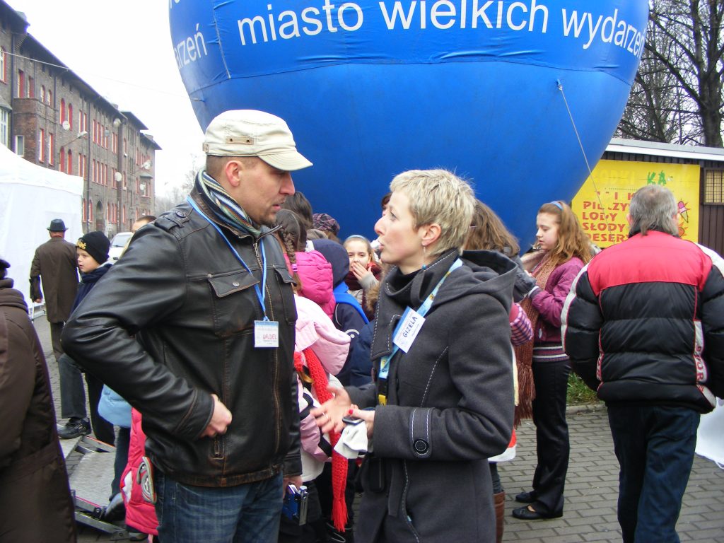 Dwoje organizator&oacute;w w trakcie rozmowy na tle innych os&oacute;b i dużego niebieskiego balonu z napisem "miasto wielkich wydarzeń".