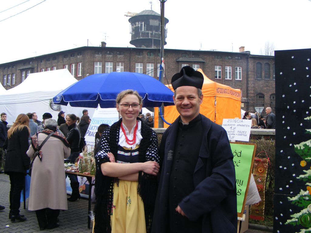 Młoda kobieta w stroju ludowym stoi obok księdza. W tle kolorowe namioty i ceglany budynek.