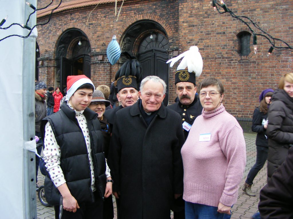 Grupa os&oacute;b pozuje do zdjęcia na tle ceglanego budynku. W centrum stoi ksiądz, obok kobieta w r&oacute;żowym swetrze, za nimi dwaj g&oacute;rnicy w galowym stroju. Po lewej młoda osoba w czapce mikołaja. W tle spacerują ludzie.