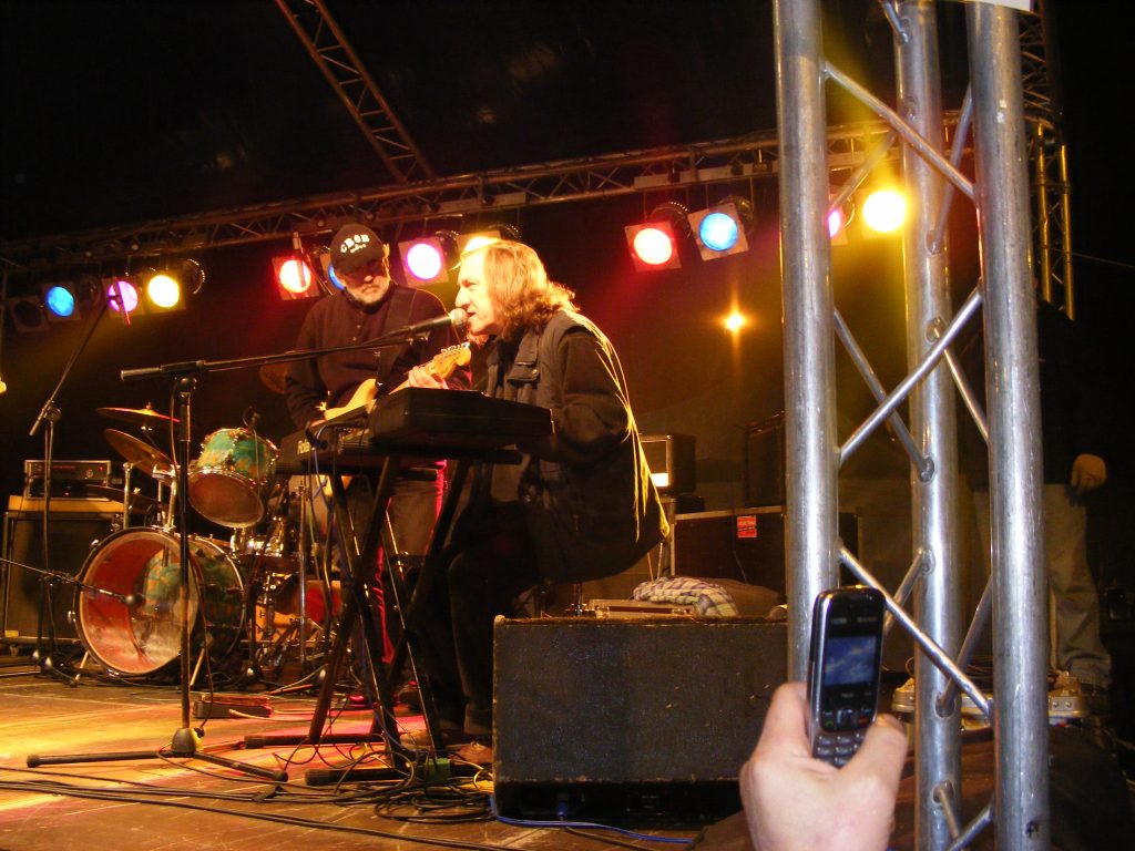 Widok na scenę, jeden mężczyzna gra na gitarze, drugi gra na klawiszach i śpiewa. W tle perkusja i kolorowe oświetlenie.