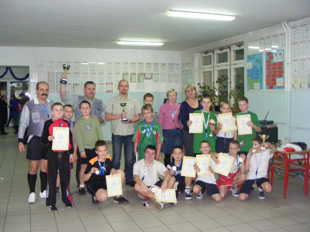 Grupowe zdjęcie dorosłych i dzieci w hali sportowej po zawodach. Uczestnicy trzymają dyplomy, puchary i medale na tle ściany z licznymi ogłoszeniami.