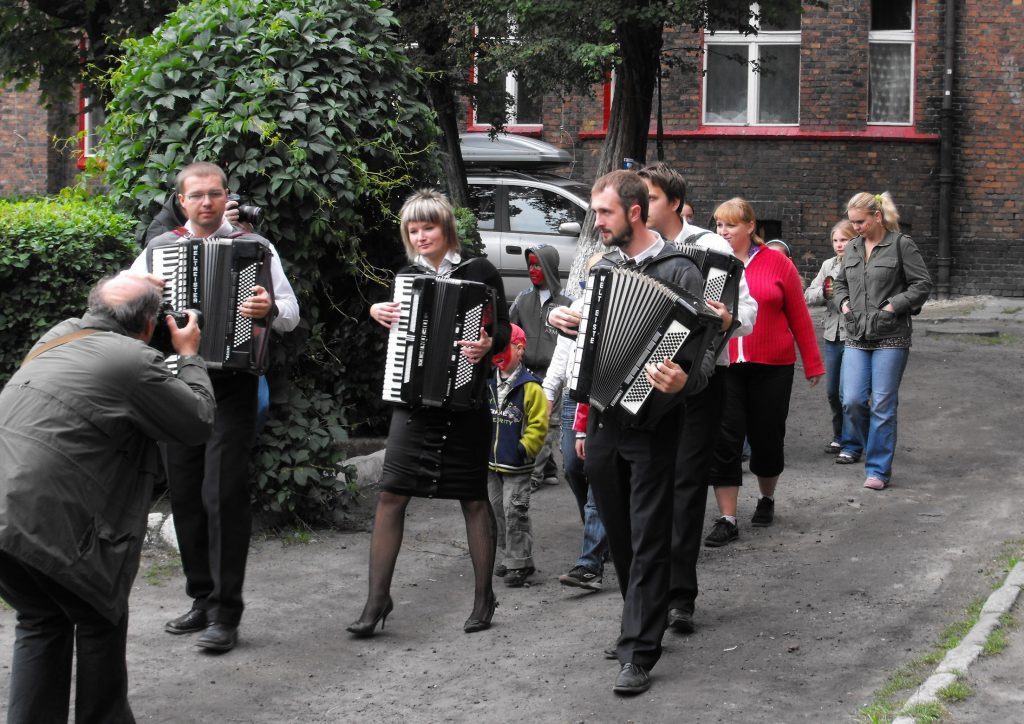 Czworo osoby grając na akordeonach, idzie drogą pomiędzy kamienicami. Fotograf robi im zdjęcie. W tle idą inne osoby.