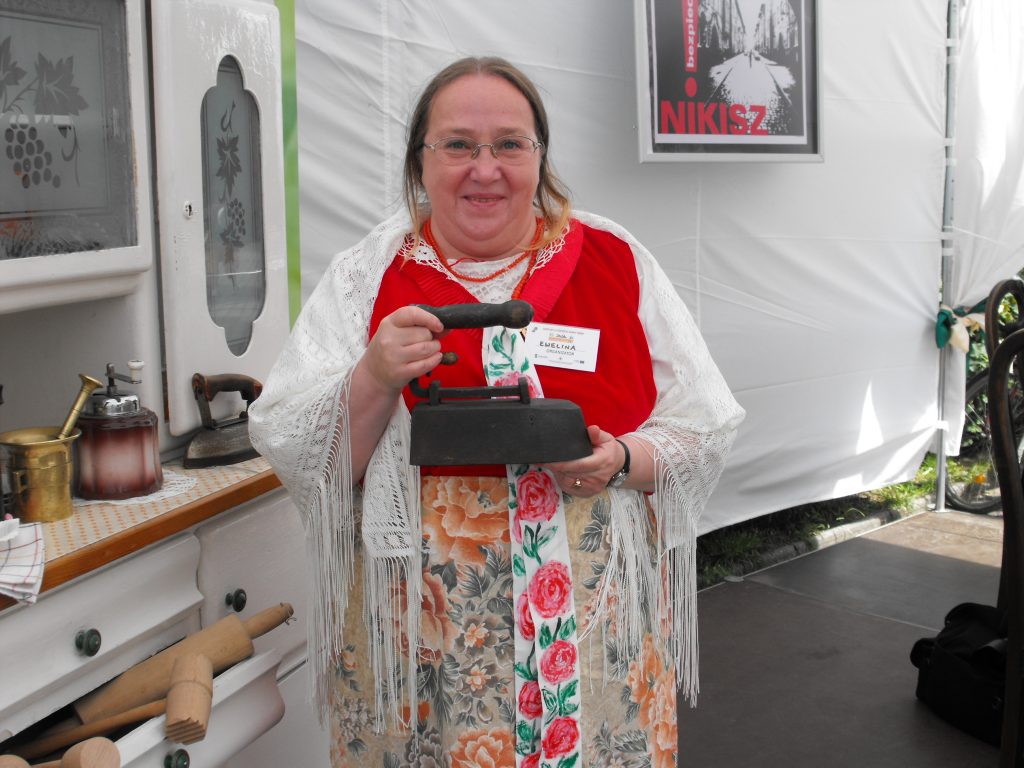 Uśmiechnięta kobieta w śląskim stroju ludowym trzyma zabytkowe żelazko z duszą. Stoi na tle białego namiotu, obok kredensu z tradycyjnymi sprzętami kuchennymi.