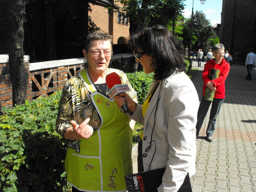 Kobieta w ż&oacute;łtym fartuchu udziela wywiadu kobiecie z mikrofonem polskiego radia Katowice. Stoją na chodniku.