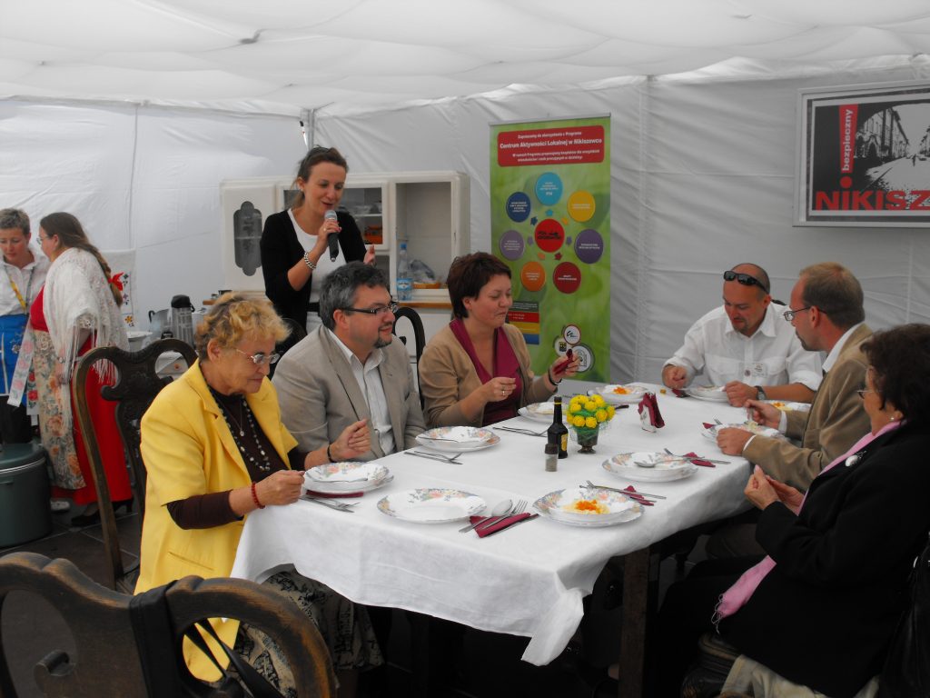 Sześć os&oacute;b siedzi przy stole pod białym, plenerowym namiotem. W tle stoi kobieta z mikrofonem w ręku.
