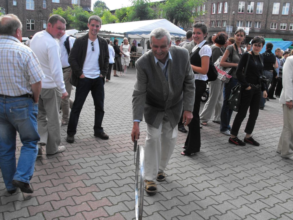 Starszy mężczyzna toczy metalową obręcz po bruku. W tle przyglądające się osoby.