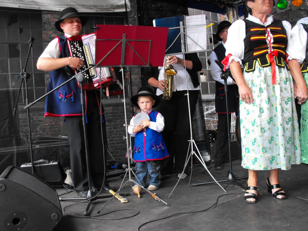 Zesp&oacute;ł ludowy występuje na scenie. Po lewej mężczyzna gra na akordeonie, obok niego stoi mały chłopiec w ludowym stroju z tamburynem, a po prawej widać fragmenty postaci w tradycyjnych strojach śląskich.