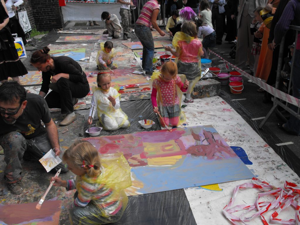 Grupa dzieci ubranych w foliowe płaszcze maluje na dużych arkuszach papieru rozłożonych na ziemi podczas plenerowych warsztat&oacute;w plastycznych.