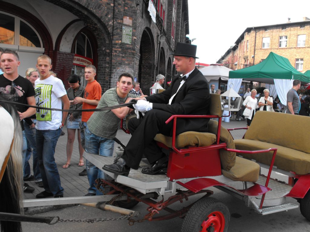 Mężczyzna w czarnym smokingu i cylindrze powozi czerwoną dorożkę. O dorożkę opiera się młody chłopak. W tle spacerujące osoby.