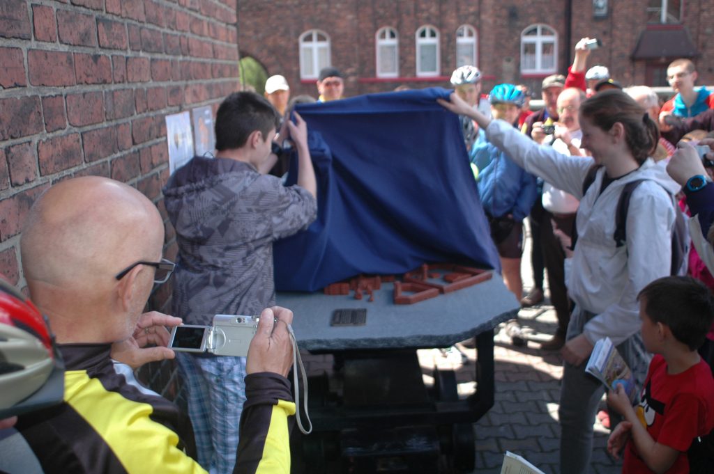 Dwie osoby podnoszą niebieską tkaninę i odsłaniają makietę osiedla Nikiszowiec. W tle grupa os&oacute;b w strojach rowerowych i kaskach.