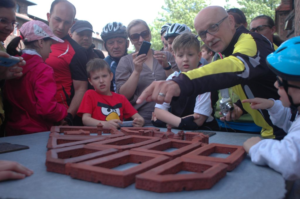 Mężczyzna w okularach opowiada i wskazuje palcem na makietę. Grupa os&oacute;b słucha i robi zdjęcia.