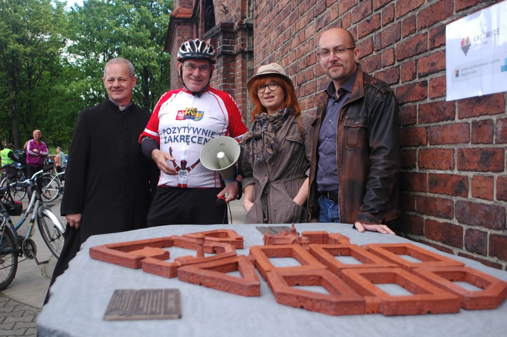 Cztery osoby w tym ksiądz, osoba w stroju rowerowym i kasku z megafonem stoi przed makietą osiedla Nikiszowiec.