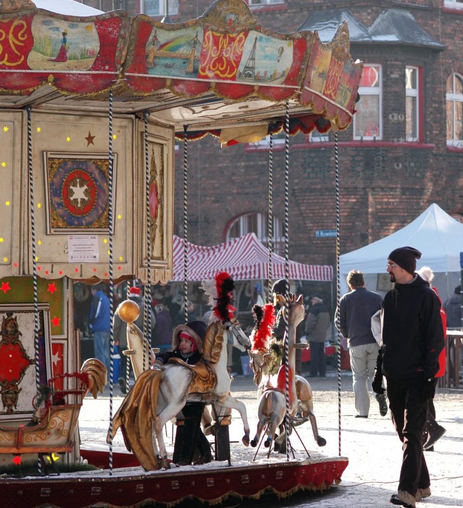 Małe dziecko przy koniku zabytkowej karuzeli. Obok stoi mężczyzna. W tle spacerują inni ludzie.