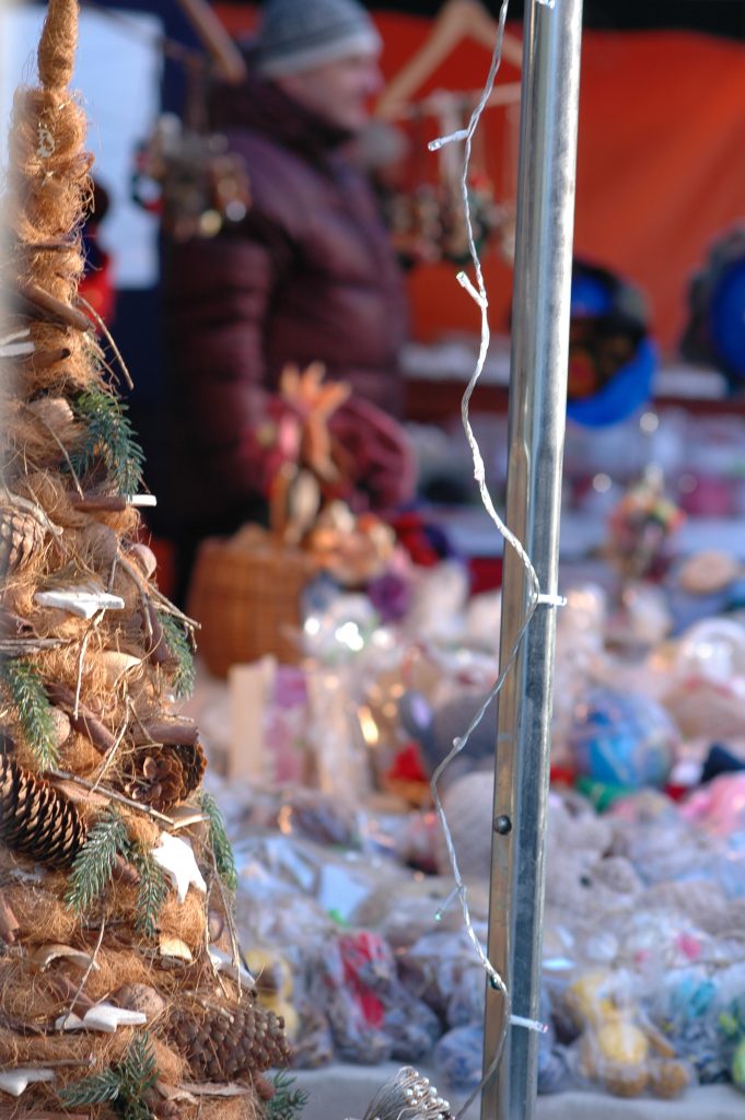 Stoisko bożonarodzeniowe z ręcznie robioną choinką z juty i szyszek na tle jarmarcznych ozd&oacute;b i światełek.