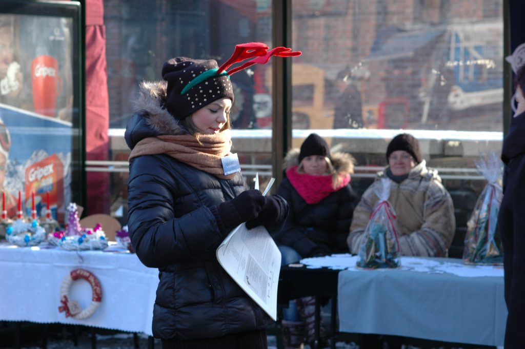 Kobieta z pluszowymi rogami renifera trzyma w rękach ulotki, w tle dwie kobiety siedzą za stołem ze świątecznymi ozdobami.