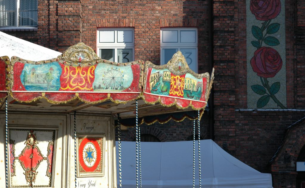 Zabytkowa karuzela z malowidłami na zadaszeniu na tle ceglanego budynku z mozaiką r&oacute;ż.