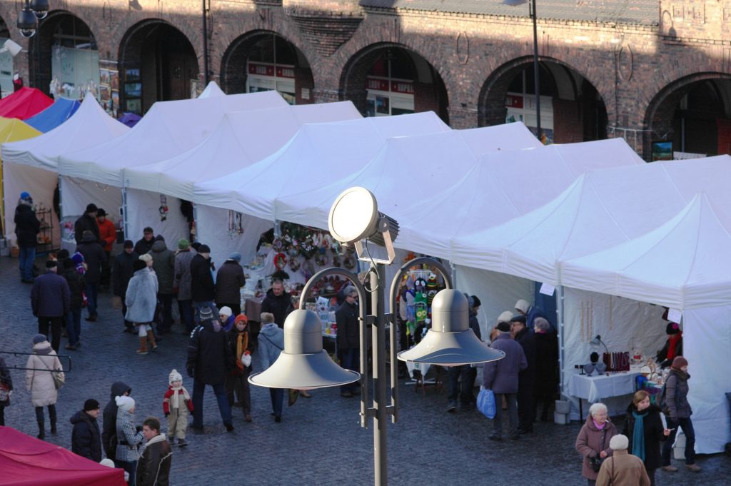 Widok z g&oacute;ry na białe namioty stoisk jarmarku. Pomiędzy spacerują ludzie.