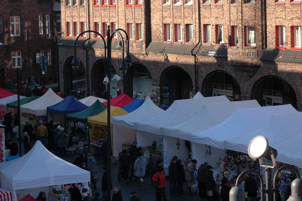 Widok z g&oacute;ry na stoiska wystawowe pod kolorowymi i białymi namiotami. Pomiędzy stoiskami przechodzą ludzie.