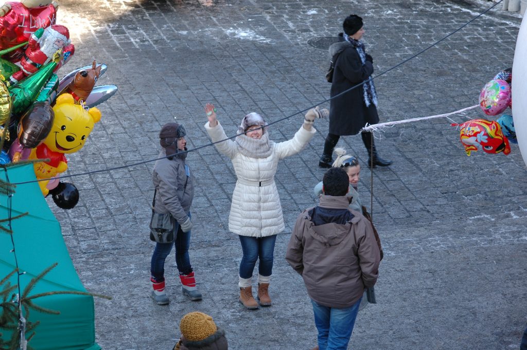 Zdjęcie z g&oacute;ry, uśmiechnięta kobieta w białej kurtce z uniesionymi rękami na tle bruku i kolorowych balon&oacute;w.
