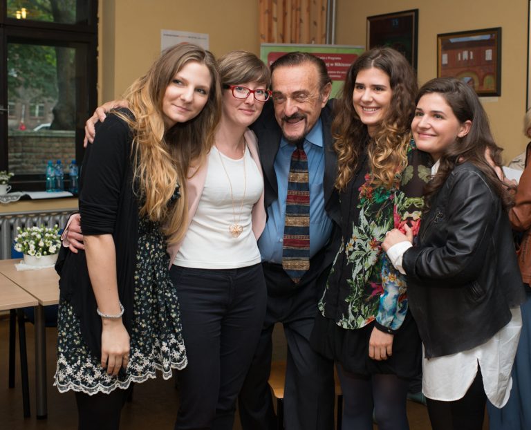 Uśmiechnięty profesor Zimbardo w ciemnym garniturze i wzorzystym krawacie obejmuje cztery młode, uśmiechnięte kobiety. Stoją w jasnym pomieszczeniu.