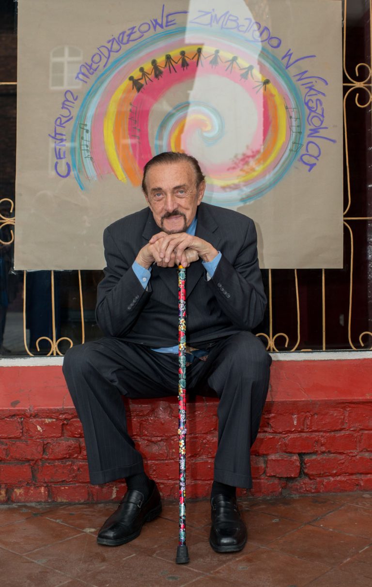 Profesor Zimbardo siedzi na czerwonym murku i opiera się o kolorową laskę. W tle plakat z tęczą i napisem centrum młodzieżowe zimbardo w Nikiszowcu.