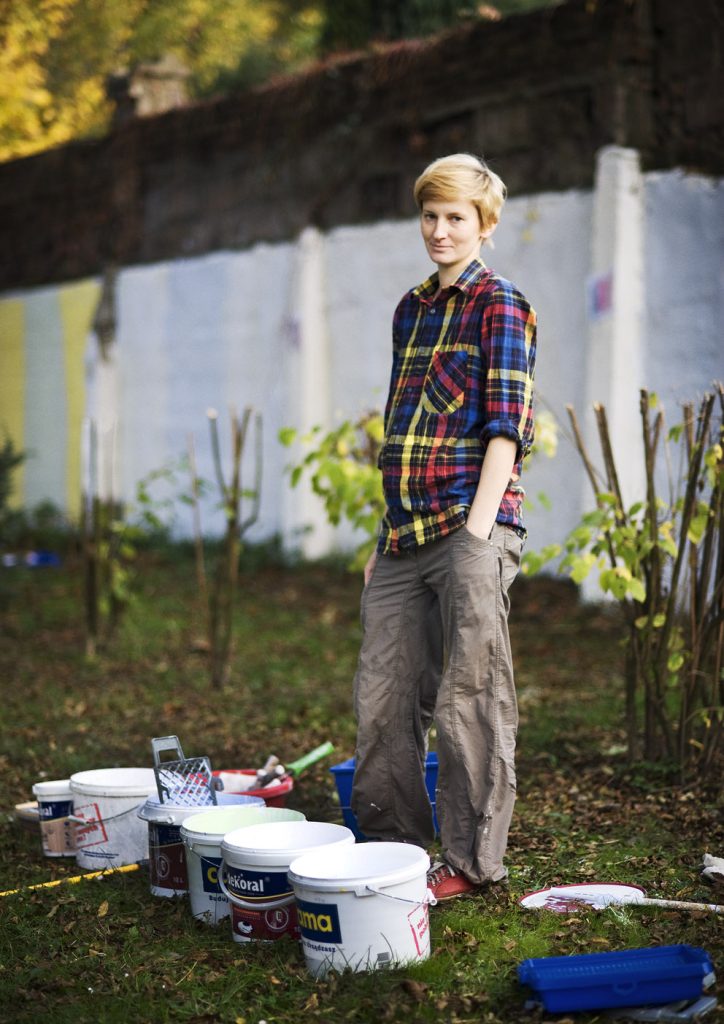 Osoba w kolorowej koszuli w kratę stoi przed wiadrami z białą farbą. W tle mur pomalowany do połowy na biało.