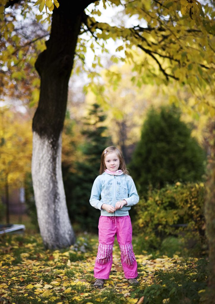 Dziewczynka w niebieskiej bluzie i r&oacute;żowych spodniach stoi pod jesiennym drzewem.