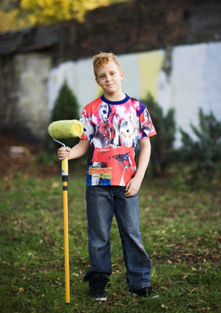 Uśmiechnięty, młody chłopak w kolorowej koszulce trzyma wałek do malowania z długim kijem. W tle fragment muru pomalowanego na biało.