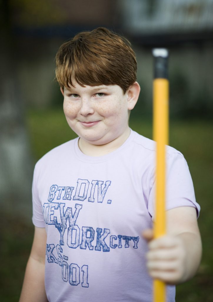 Młody, uśmiechnięty chłopak trzyma przed sobą ż&oacute;łty kij od wałka do malowania.