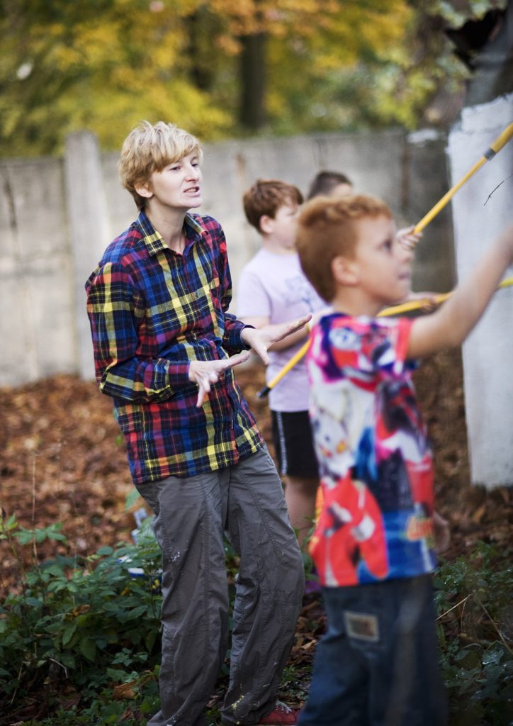 Osoba w kolorowej koszuli w kratę m&oacute;wi do grupy dzieci, kt&oacute;re trzymają w rękach ż&oacute;łte kije od wałk&oacute;w do malowania.