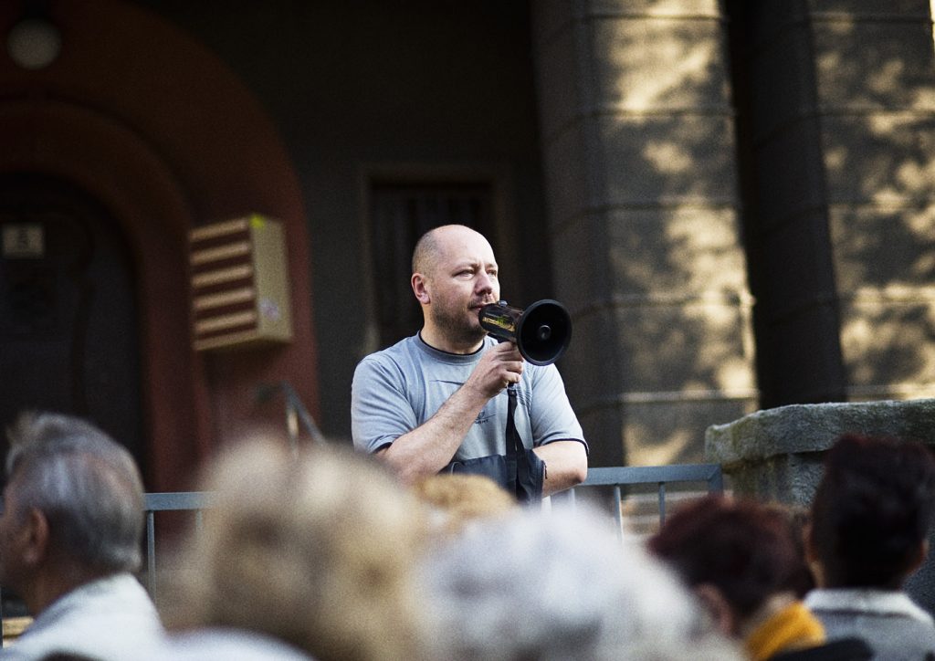 Mężczyzna w szarej koszulce m&oacute;wi do megafonu. W tle fragment ciemnej elewacji budynku.