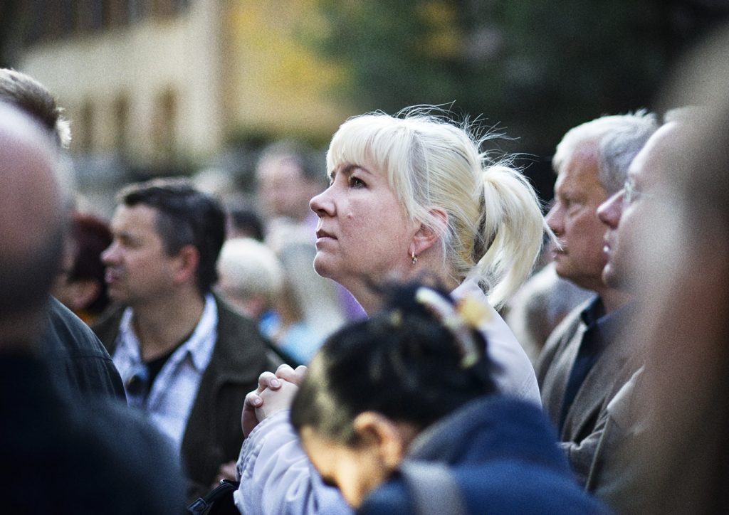 kobieta z jasnymi włosami zebranymi w kucyk spogląda z zaciekawieniem. W koło niej rozmyte postacie.