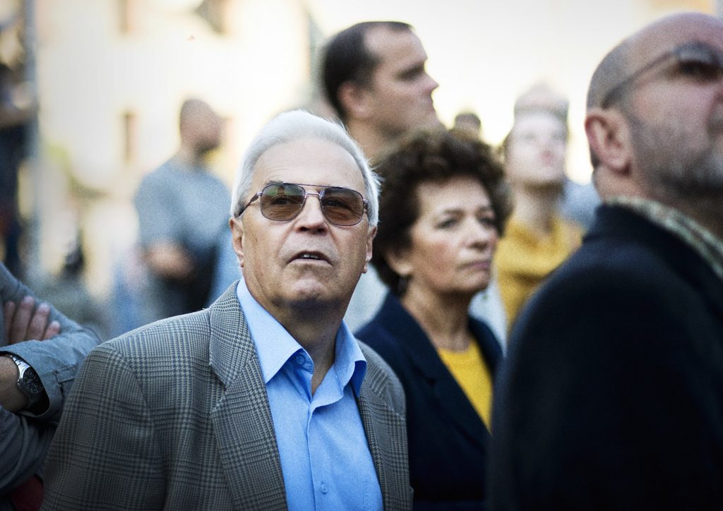 Starszy mężczyzna w kraciastej marynarce i okularach przeciwsłonecznych spogląda przed siebie. W tle rozmyte postacie.