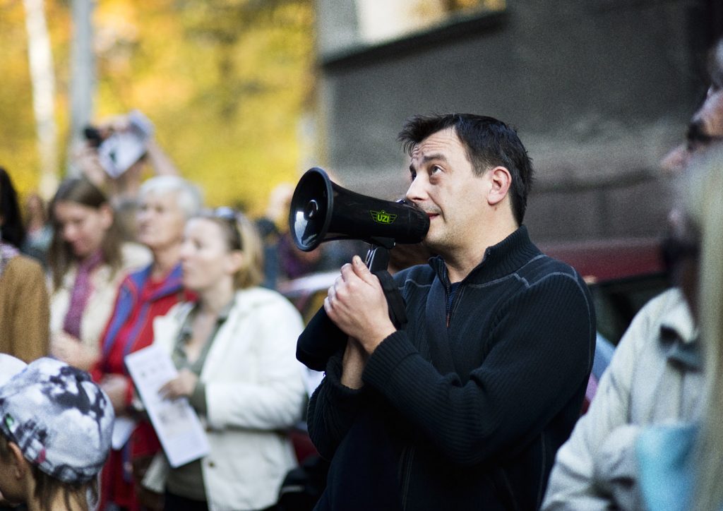 Mężczyzna w czarnym polarze m&oacute;wi do megafonu spoglądając w g&oacute;rę. Stoi na tle grupy os&oacute;b przed budynkiem.