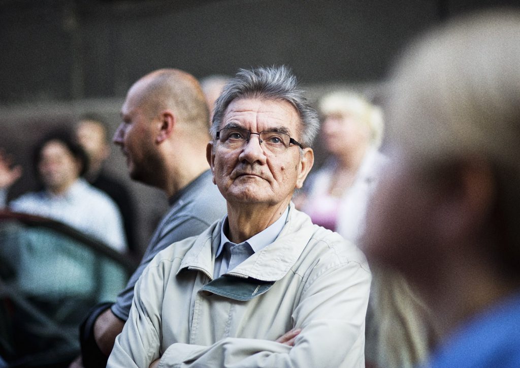 Starszy mężczyzna w jasnej kurtce i okularach spogląda w g&oacute;rę. W tle kilka rozmytych postaci.