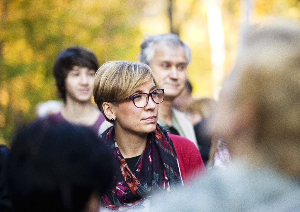 Kobieta w okularach i wzorzystej apaszce patrzy w lewo. W tle rozmyte postaci ludzi i jesienne drzewa.